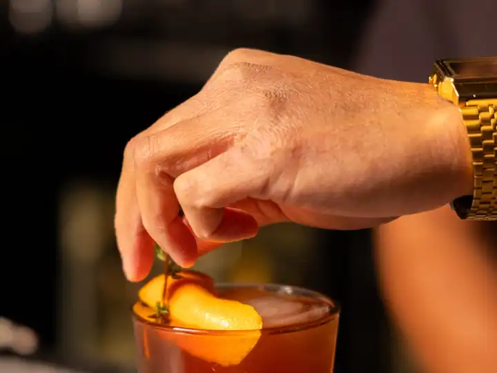 Hand twisting an orange peel garnish over a dark amber cocktail in a rocks glass