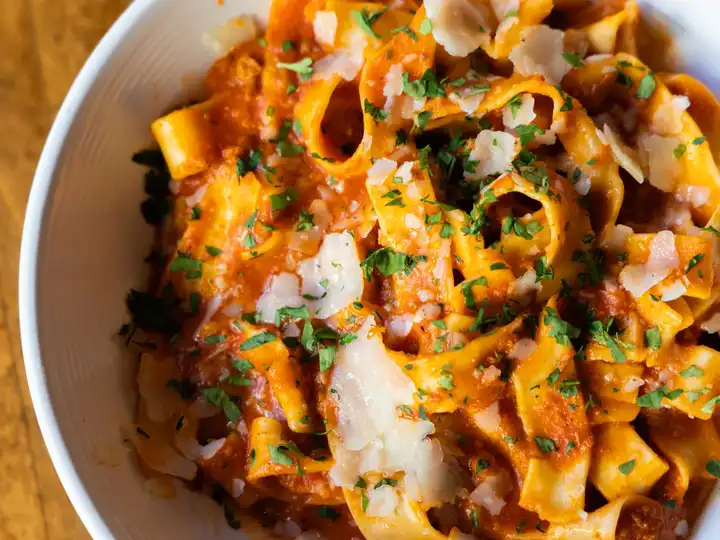 Close-up of ribbon pasta in red sauce topped with shaved cheese and chopped herbs in a white bowl