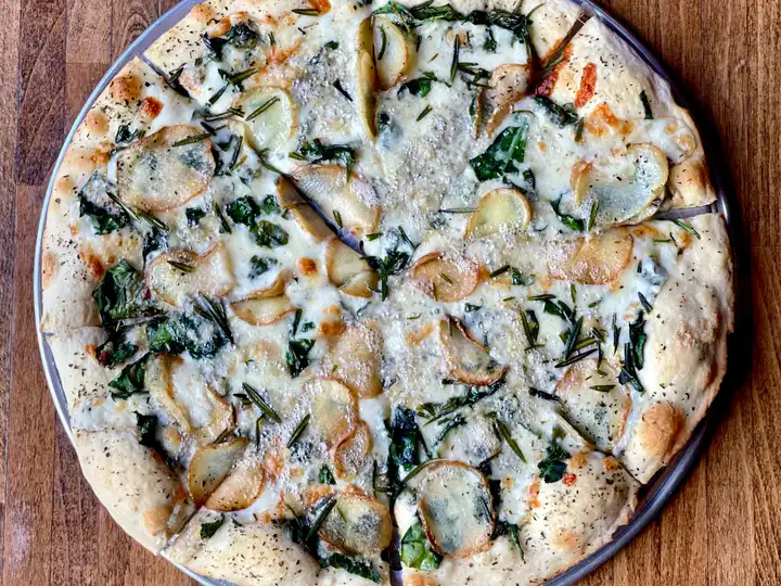 Round pizza on a metal pan topped with white cheese, sliced potatoes, and rosemary sprigs
