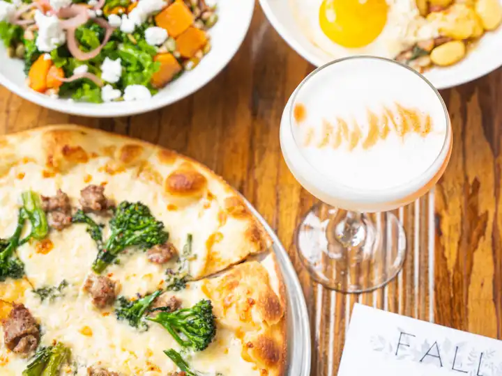Pizza with sausage, side salad, bowl with a fried egg, and a foam-topped cocktail
